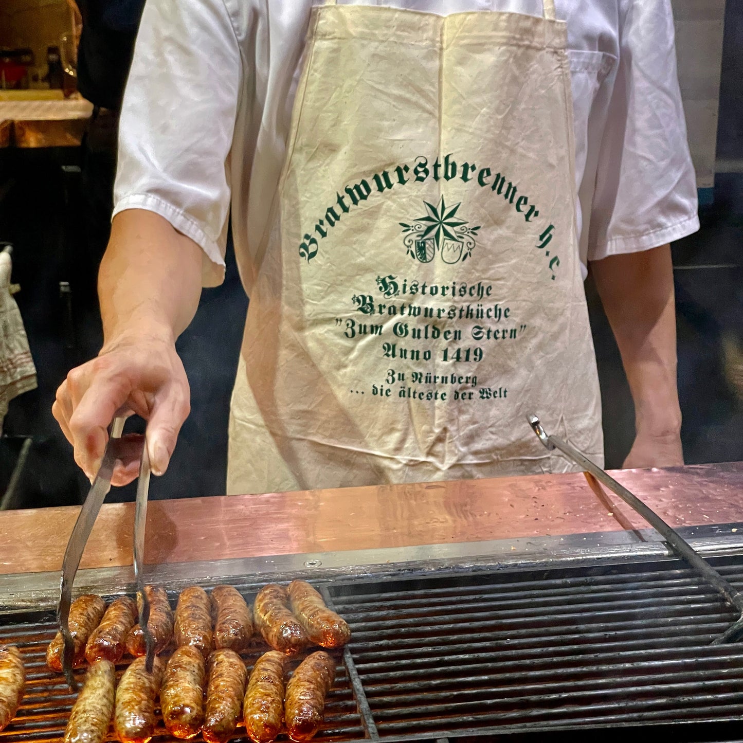 Ein Koch in einer naturfarbenen Leinen-Schürze mit der Aufschrift "Bratwurstbrenner h.c." steht an einem Grill und wendet mit einer Zange mehrere Nürnberger Rostbratwürste. Die Schürze trägt den historischen Aufdruck der Bratwurstküche "Zum Gulden Stern", der ältesten Bratwurstküche der Welt. Die Szene ist von aufsteigendem Rauch umgeben, was die traditionelle Zubereitung über offenem Feuer betont. Im Hintergrund ist die rustikale Atmosphäre der historischen Wirtshausküche zu erkennen.