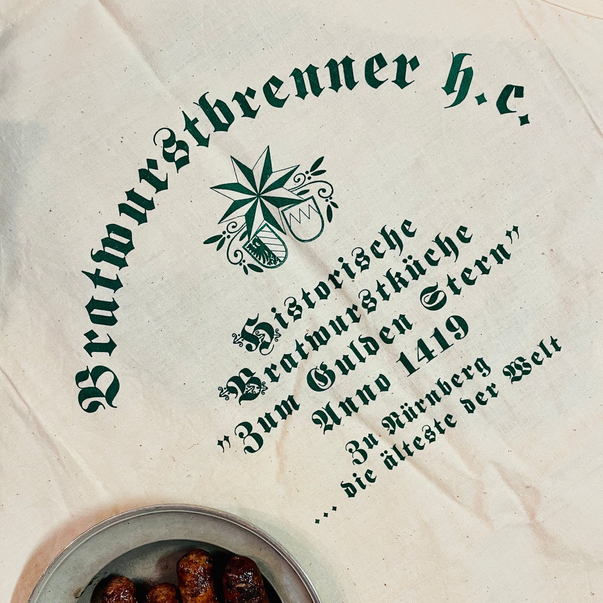 Eine naturfarbene Schürze aus Leinen mit historischem, grünen Aufdruck liegt auf einem Tisch. Der Schriftzug "Bratwurstbrenner h.c." sowie die Inschrift "Historische Bratwurstküche Zum Gulden Stern Anno 1419 – die älteste der Welt" sind deutlich zu sehen. Ein runder Zinnteller mit vier gegrillten Nürnberger Bratwürsten ergänzt das rustikale Ambiente und betont die kulinarische Tradition der Schürze.