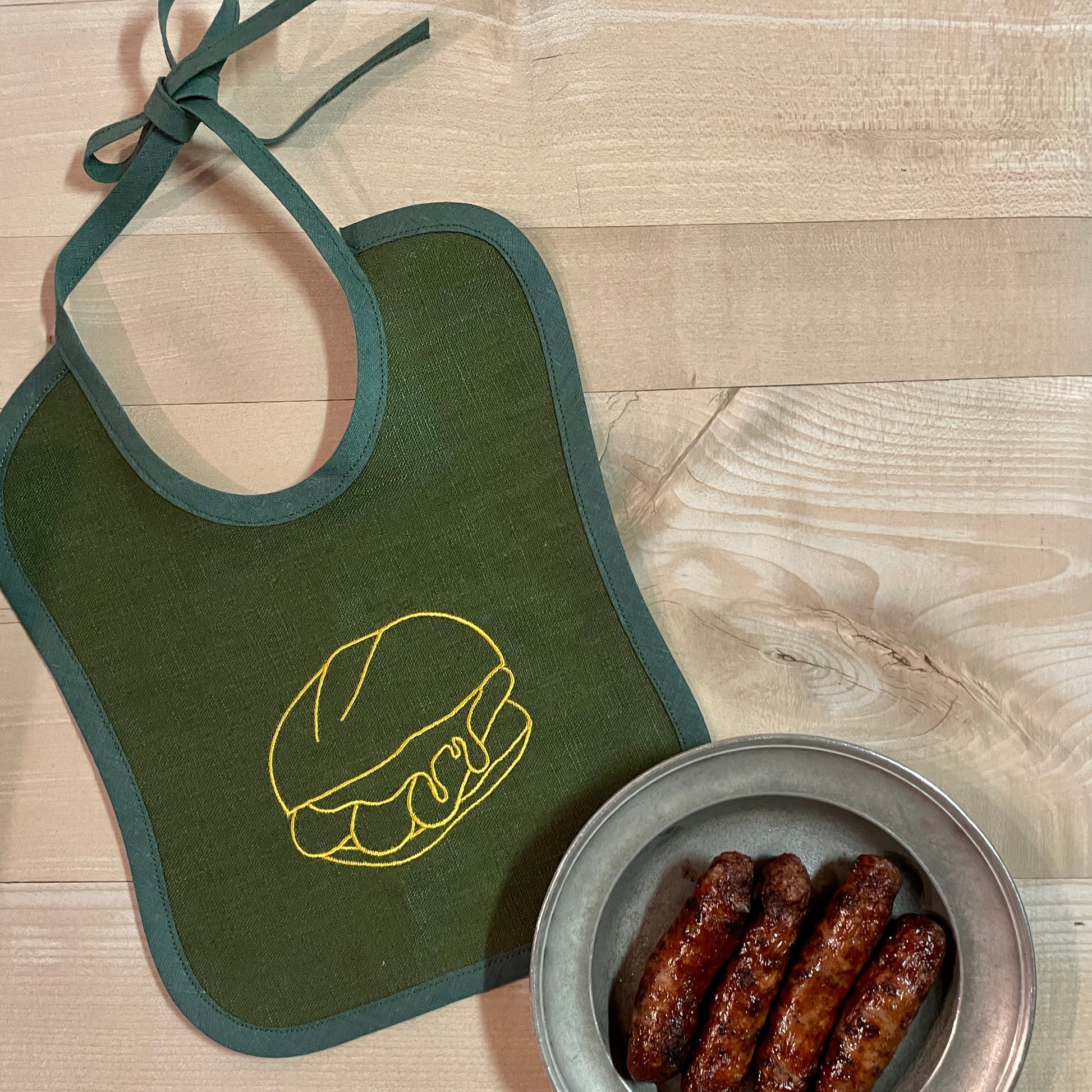 Ein grünes Baby-Lätzchen aus Leinen mit goldgelber Stickerei eines Bratwurstbrötchens liegt auf einem hellen Holztisch. Das Lätzchen hat eine dunkelgrüne Einfassung und lange Stoffbänder zum Binden. Daneben steht ein runder Zinnteller mit vier gegrillten Bratwürsten, die das fränkische Flair des Motivs unterstreichen. Die Szene strahlt eine rustikale, gemütliche Atmosphäre aus und hebt das hochwertige Material sowie das liebevolle Design des Baby-Lätzchens hervor.