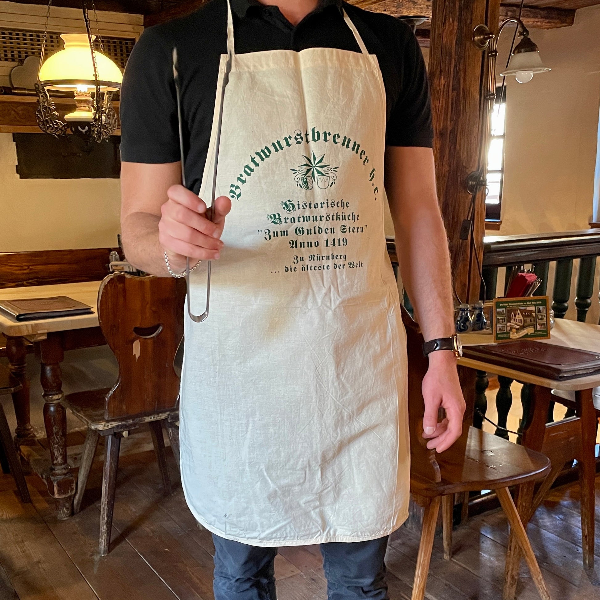 Ein Mann trägt eine naturfarbene Leinen-Schürze mit dem Aufdruck "Bratwurstbrenner h.c." und der historischen Inschrift der Bratwurstküche "Zum Gulden Stern", der ältesten Bratwurstküche der Welt. Er hält eine Grillzange in der Hand und steht in einem rustikalen, traditionellen Wirtshaus mit Holzmöbeln und gemütlicher Beleuchtung. Die Schürze hat eine klassische Form mit Nackenband und langen Bändern zum Binden. Die Szene strahlt eine authentische fränkische Wirtshaus-Atmosphäre aus.
