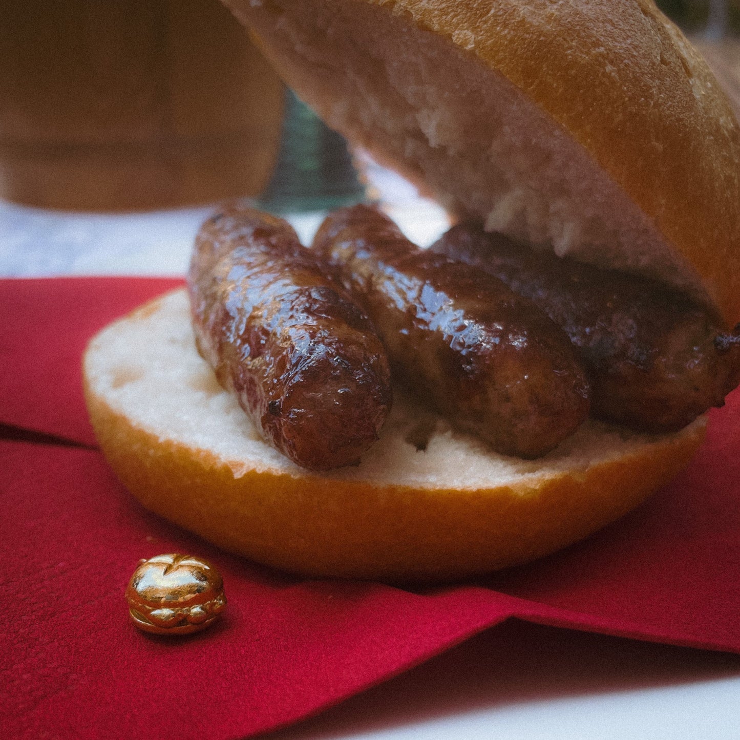 Ein klassisches fränkisches „3 im Weggla“ – drei saftige Nürnberger Rostbratwürste in einem knusprigen Brötchen – serviert auf einem weißen Teller mit einer roten Serviette. Daneben liegt ein filigraner, vergoldeter Charm in Form des ikonischen „3 im Weggla“-Motivs, der die kulinarische Tradition Nürnbergs als edles Schmuckstück widerspiegelt. Im Hintergrund ist ein traditioneller Bierkrug zu erkennen, der die gemütliche Wirtshaus-Atmosphäre unterstreicht.