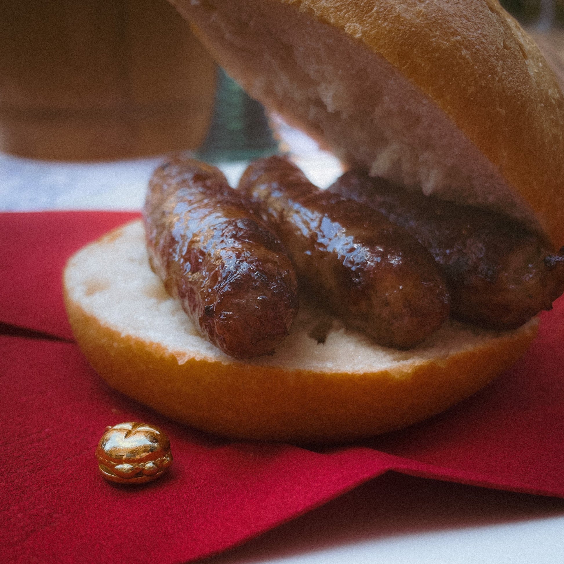 Ein klassisches fränkisches „3 im Weggla“ – drei saftige Nürnberger Rostbratwürste in einem knusprigen Brötchen – serviert auf einem weißen Teller mit einer roten Serviette. Daneben liegt ein filigraner, vergoldeter Charm in Form des ikonischen „3 im Weggla“-Motivs, der die kulinarische Tradition Nürnbergs als edles Schmuckstück widerspiegelt. Im Hintergrund ist ein traditioneller Bierkrug zu erkennen, der die gemütliche Wirtshaus-Atmosphäre unterstreicht.