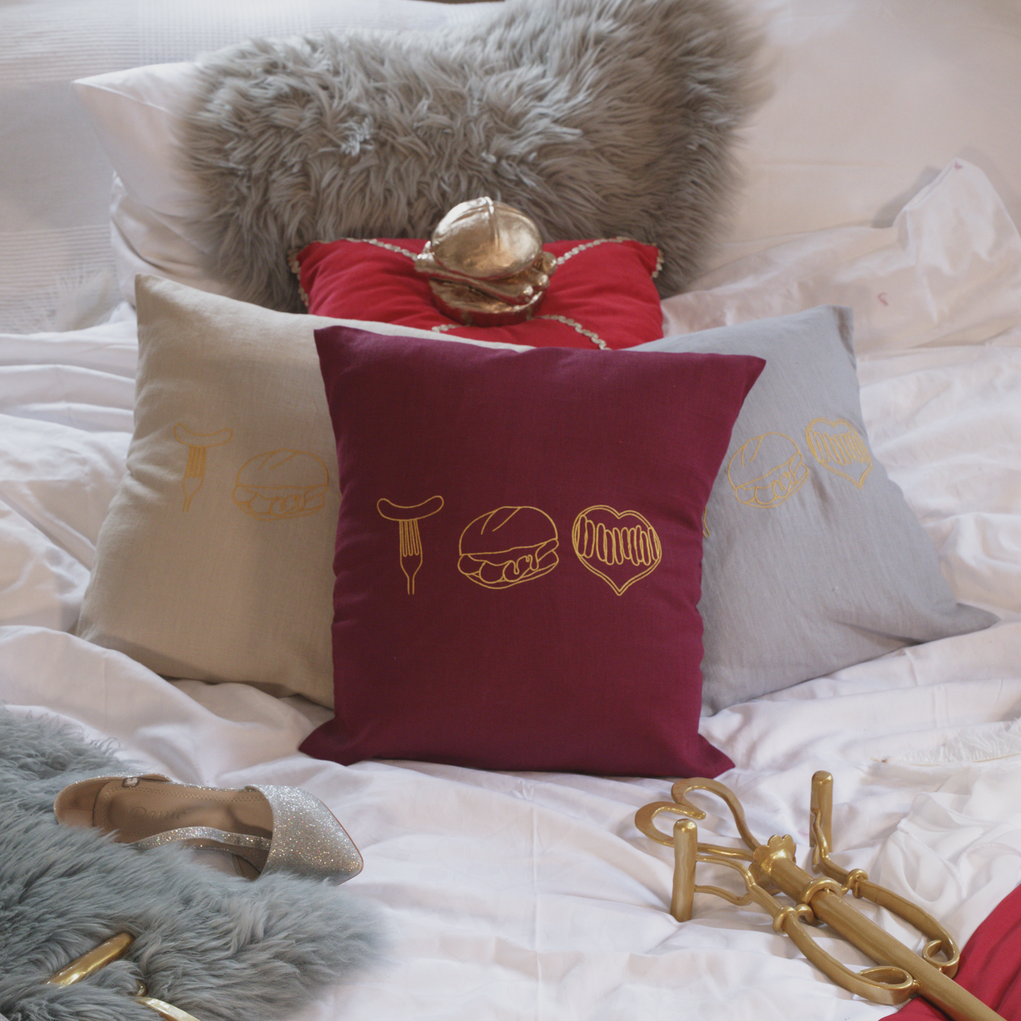 Mehrere dekorative Leinenkissen in den Farben Mondgrau, Sandstein und Weinrot auf einem gemütlich gestalteten Bett. Die Kissen sind mit goldenen Stickereien versehen, die fränkische Bratwurst-Motive zeigen: eine Bratwurst auf der Gabel, drei Bratwürste im Brötchen und ein Bratwurst-Herz. Im Hintergrund liegen ein graues Kunstfell-Kissen, ein rotes Samtkissen mit einem goldenen Detail sowie stilvolle Accessoires wie ein silberner Schuh und eine goldene Zepter.