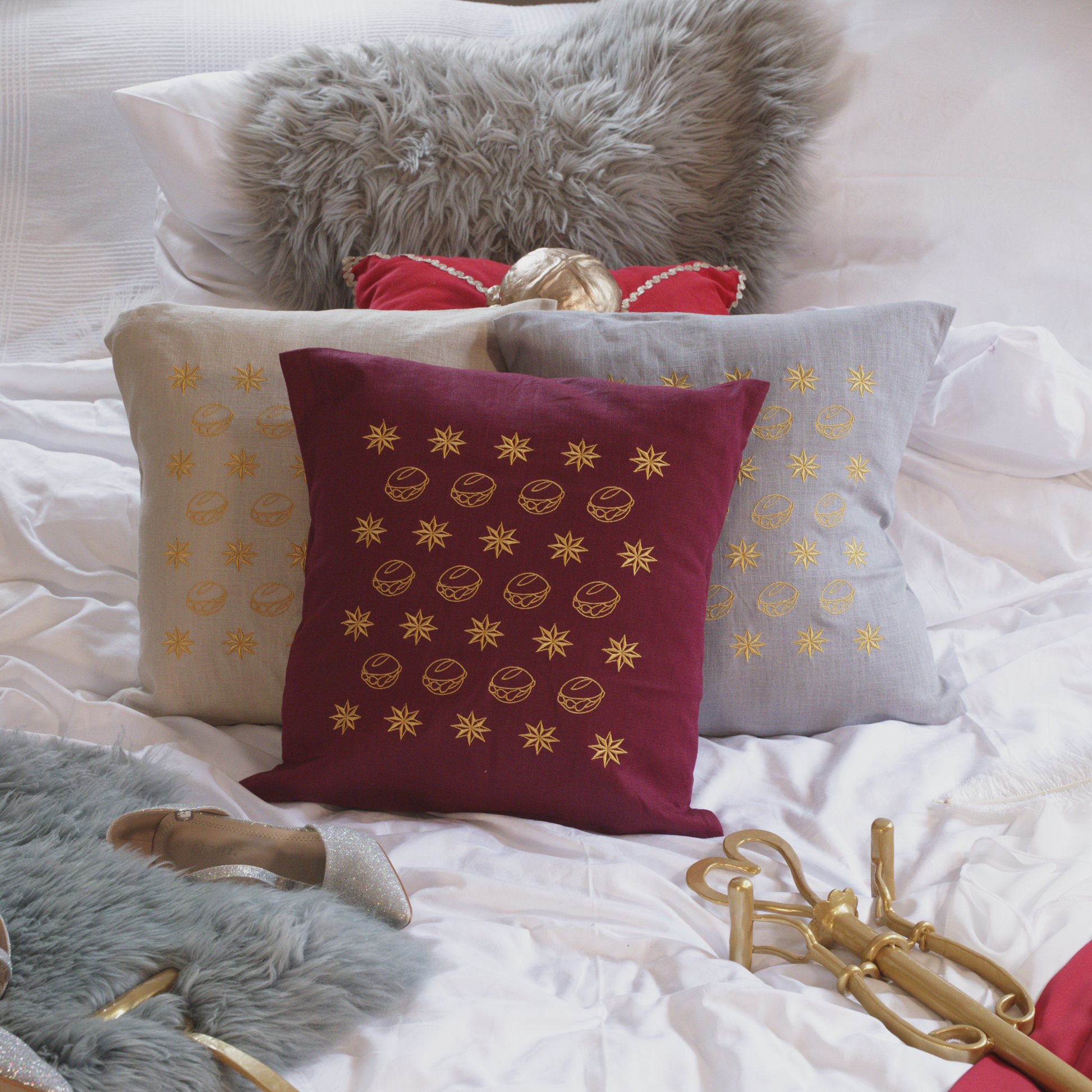 Drei dekorative Leinenkissen in den Farben Mondgrau, Sandstein und Weinrot mit goldener Stickerei, die eine Musterung aus kleinen Brötchen („Weggla“) und Sternen („Sternla“) zeigt. Die Kissen sind inklusive Füllung und liegen auf einem stilvoll arrangierten Bett mit weiteren Dekoelementen wie einem grauen Kunstfell-Kissen, einem roten Samtkissen mit goldenen Details, einem silberne Schuh und einer goldenem Zepter.