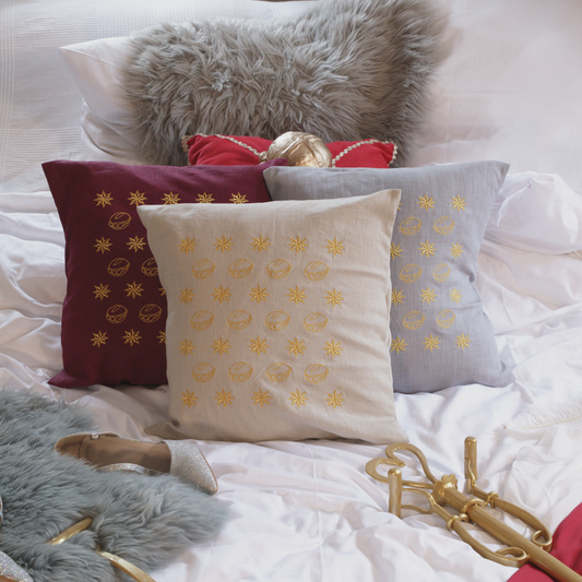 Drei dekorative Leinenkissen in den Farben Mondgrau, Sandstein und Weinrot mit goldener Stickerei, die eine Musterung aus kleinen Brötchen („Weggla“) und Sternen („Sternla“) zeigt. Die Kissen sind inklusive Füllung und liegen auf einem stilvoll arrangierten Bett mit weiteren Dekoelementen wie einem grauen Kunstfell-Kissen, einem roten Samtkissen mit goldenen Details, einem silberne Schuh und einer goldenem Zepter.