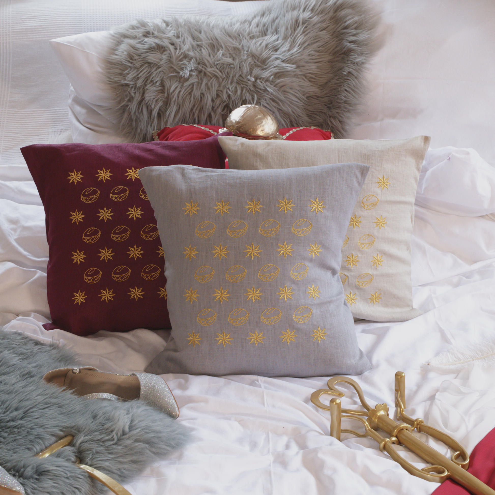 Drei dekorative Leinenkissen in den Farben Mondgrau, Sandstein und Weinrot mit goldener Stickerei, die eine Musterung aus kleinen Brötchen („Weggla“) und Sternen („Sternla“) zeigt. Die Kissen sind inklusive Füllung und liegen auf einem stilvoll arrangierten Bett mit weiteren Dekoelementen wie einem grauen Kunstfell-Kissen, einem roten Samtkissen mit goldenen Details, einem silberne Schuh und einer goldenem Zepter.
