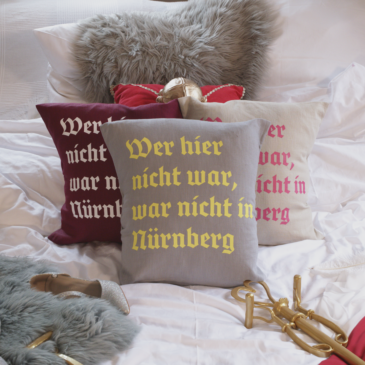 Drei dekorative Leinenkissen in den Farben Mondgrau, Sandstein und Weinrot mit gedrucktem Zitat „Wer hier nicht war, war nicht in Nürnberg“ in historischer Frakturschrift. Die Kissen sind inklusive Füllung und liegen auf einem geschmackvoll arrangierten Bett mit weiteren Dekoelementen wie einem grauen Kunstfell-Kissen, einem roten Samtkissen mit goldenen Details, einem silbernen Schuh und einem goldenen Zepter.