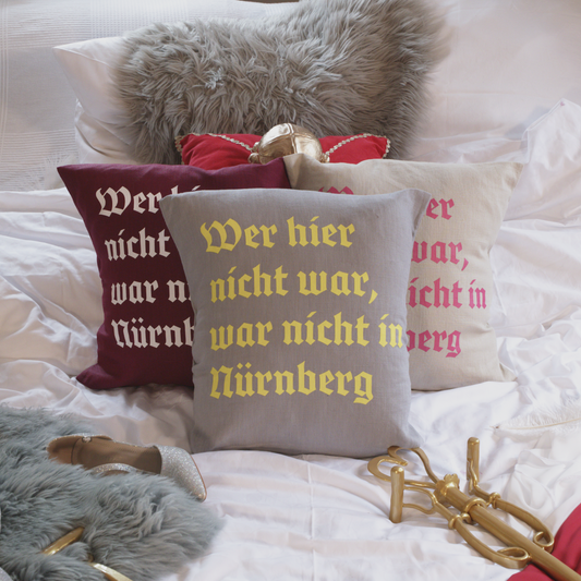Drei dekorative Leinenkissen in den Farben Mondgrau, Sandstein und Weinrot mit gedrucktem Zitat „Wer hier nicht war, war nicht in Nürnberg“ in historischer Frakturschrift. Die Kissen sind inklusive Füllung und liegen auf einem geschmackvoll arrangierten Bett mit weiteren Dekoelementen wie einem grauen Kunstfell-Kissen, einem roten Samtkissen mit goldenen Details, einem silbernen Schuh und einem goldenen Zepter.