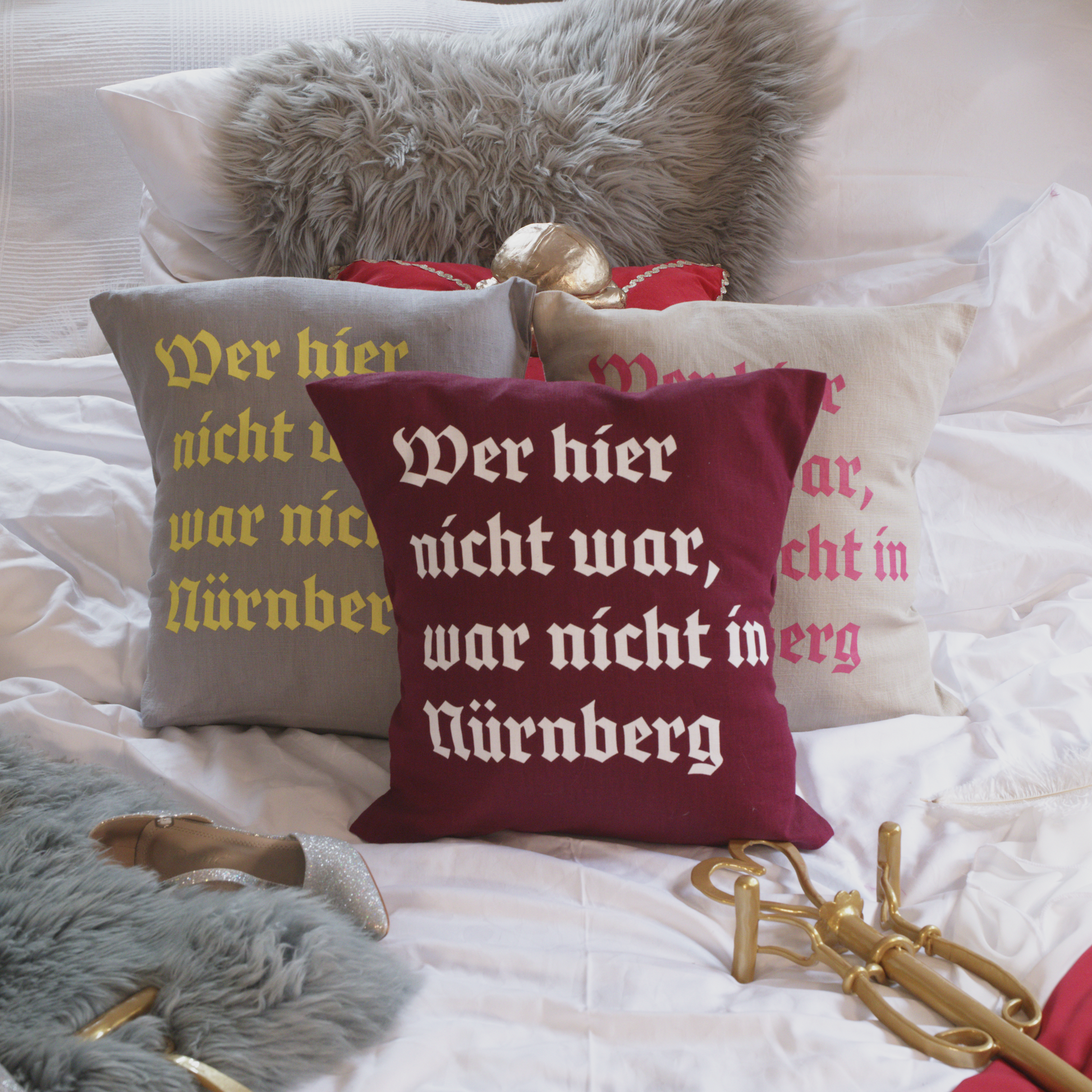 Drei dekorative Leinenkissen in den Farben Mondgrau, Sandstein und Weinrot mit gedrucktem Zitat „Wer hier nicht war, war nicht in Nürnberg“ in historischer Frakturschrift. Die Kissen sind inklusive Füllung und liegen auf einem geschmackvoll arrangierten Bett mit weiteren Dekoelementen wie einem grauen Kunstfell-Kissen, einem roten Samtkissen mit goldenen Details, einem silbernen Schuh und einem goldenen Zepter.