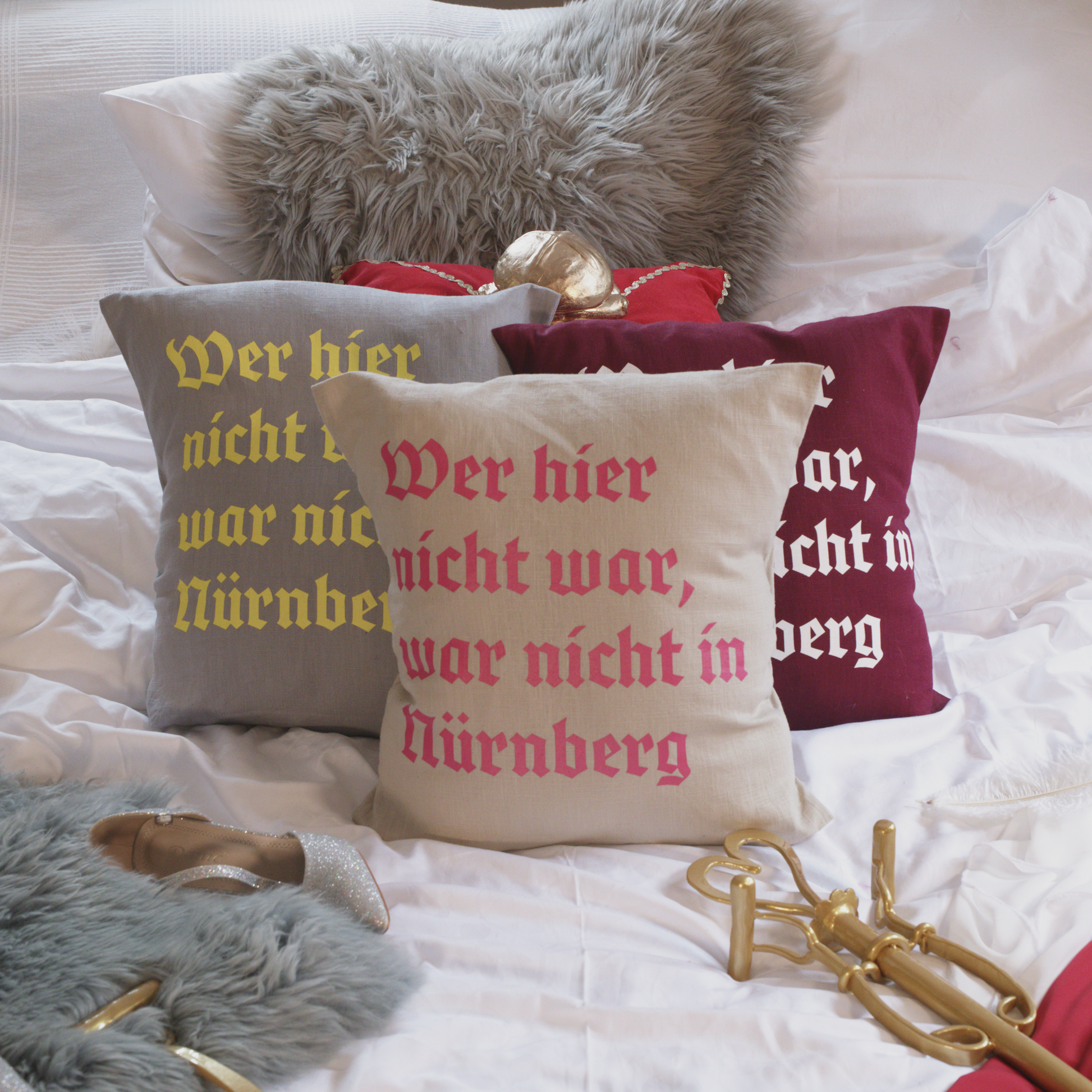 Drei dekorative Leinenkissen in den Farben Mondgrau, Sandstein und Weinrot mit gedrucktem Zitat „Wer hier nicht war, war nicht in Nürnberg“ in historischer Frakturschrift. Die Kissen sind inklusive Füllung und liegen auf einem geschmackvoll arrangierten Bett mit weiteren Dekoelementen wie einem grauen Kunstfell-Kissen, einem roten Samtkissen mit goldenen Details, einem silbernen Schuh und einem goldenen Zepter.