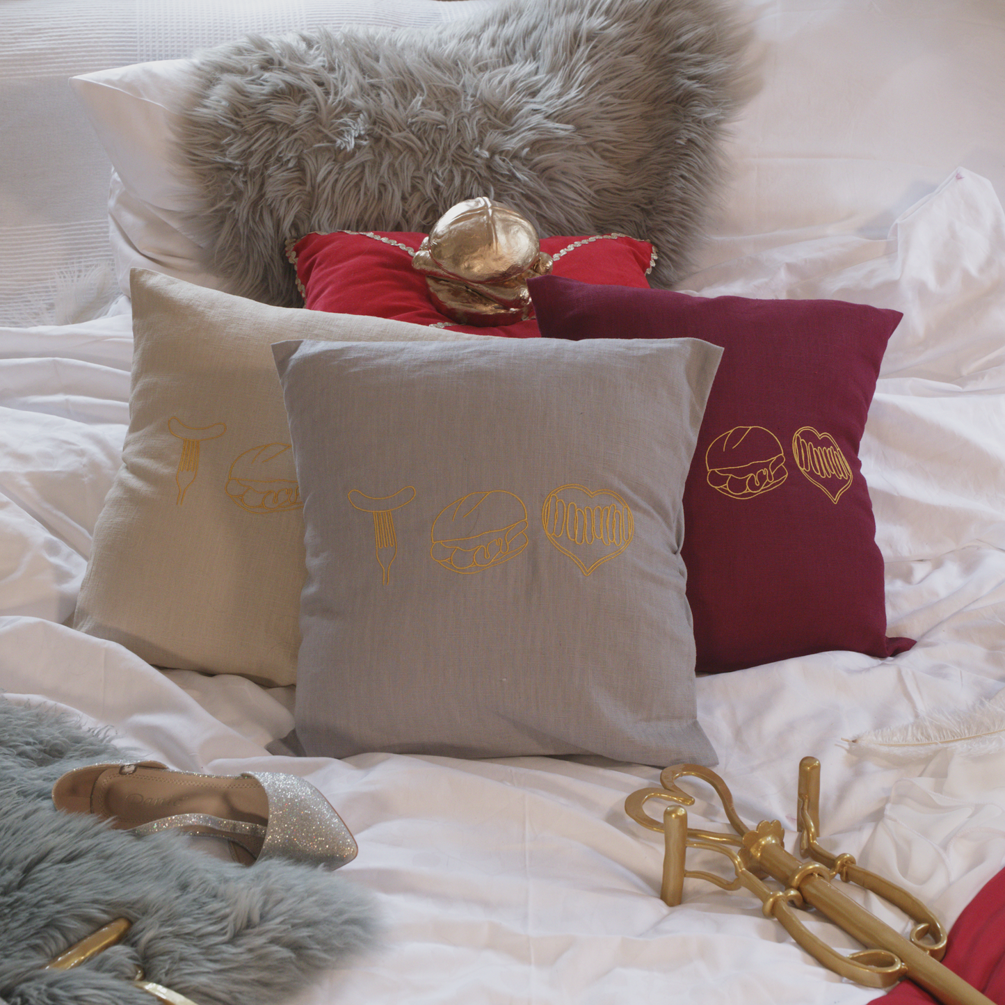 Mehrere dekorative Leinenkissen in den Farben Mondgrau, Sandstein und Weinrot auf einem gemütlich gestalteten Bett. Die Kissen sind mit goldenen Stickereien versehen, die fränkische Bratwurst-Motive zeigen: eine Bratwurst auf der Gabel, drei Bratwürste im Brötchen und ein Bratwurst-Herz. Im Hintergrund liegen ein graues Kunstfell-Kissen, ein rotes Samtkissen mit einem goldenen Detail sowie stilvolle Accessoires wie ein silberner Schuh und eine goldene Zepter.