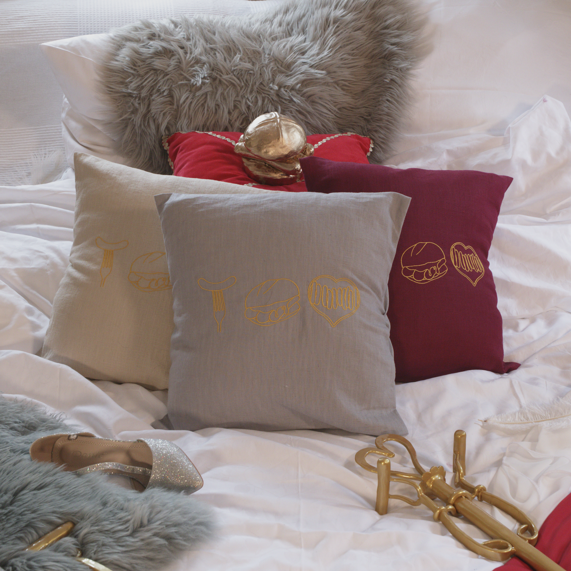 Mehrere dekorative Leinenkissen in den Farben Mondgrau, Sandstein und Weinrot auf einem gemütlich gestalteten Bett. Die Kissen sind mit goldenen Stickereien versehen, die fränkische Bratwurst-Motive zeigen: eine Bratwurst auf der Gabel, drei Bratwürste im Brötchen und ein Bratwurst-Herz. Im Hintergrund liegen ein graues Kunstfell-Kissen, ein rotes Samtkissen mit einem goldenen Detail sowie stilvolle Accessoires wie ein silberner Schuh und eine goldene Zepter.