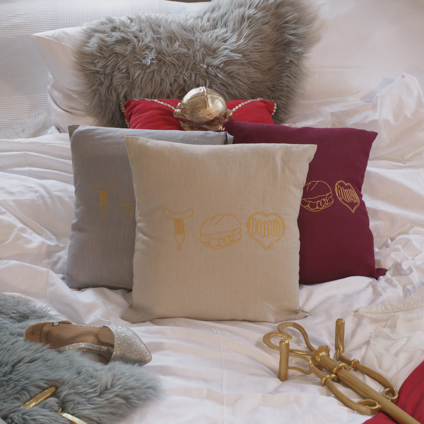 Mehrere dekorative Leinenkissen in den Farben Mondgrau, Sandstein und Weinrot auf einem gemütlich gestalteten Bett. Die Kissen sind mit goldenen Stickereien versehen, die fränkische Bratwurst-Motive zeigen: eine Bratwurst auf der Gabel, drei Bratwürste im Brötchen und ein Bratwurst-Herz. Im Hintergrund liegen ein graues Kunstfell-Kissen, ein rotes Samtkissen mit einem goldenen Detail sowie stilvolle Accessoires wie ein silberner Schuh und eine goldene Zepter.