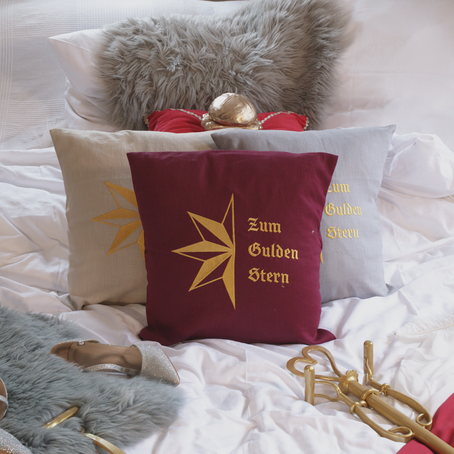 Drei dekorative Leinenkissen in den Farben Mondgrau, Sandstein und Weinrot mit goldener Stickerei, die den stilisierten Stern und die Aufschrift „Zum Gulden Stern“ zeigt. Die Kissen sind inklusive Füllung und liegen auf einem elegant arrangierten Bett mit weiteren Dekoelementen wie einem grauen Kunstfell-Kissen, einem roten Samtkissen mit goldenen Details, einem silbernen Schuh und einer goldenem Zepter.