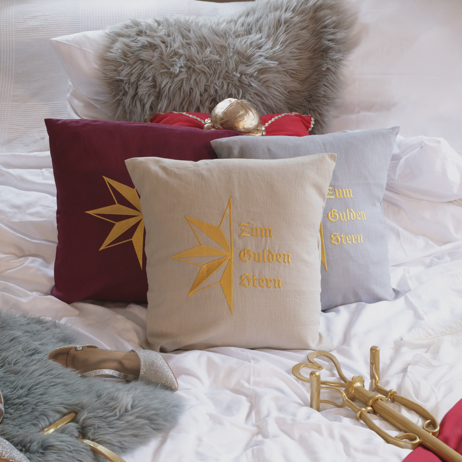 Drei dekorative Leinenkissen in den Farben Mondgrau, Sandstein und Weinrot mit goldener Stickerei, die den stilisierten Stern und die Aufschrift „Zum Gulden Stern“ zeigt. Die Kissen sind inklusive Füllung und liegen auf einem elegant arrangierten Bett mit weiteren Dekoelementen wie einem grauen Kunstfell-Kissen, einem roten Samtkissen mit goldenen Details, einem silbernen Schuh und einer goldenem Zepter.