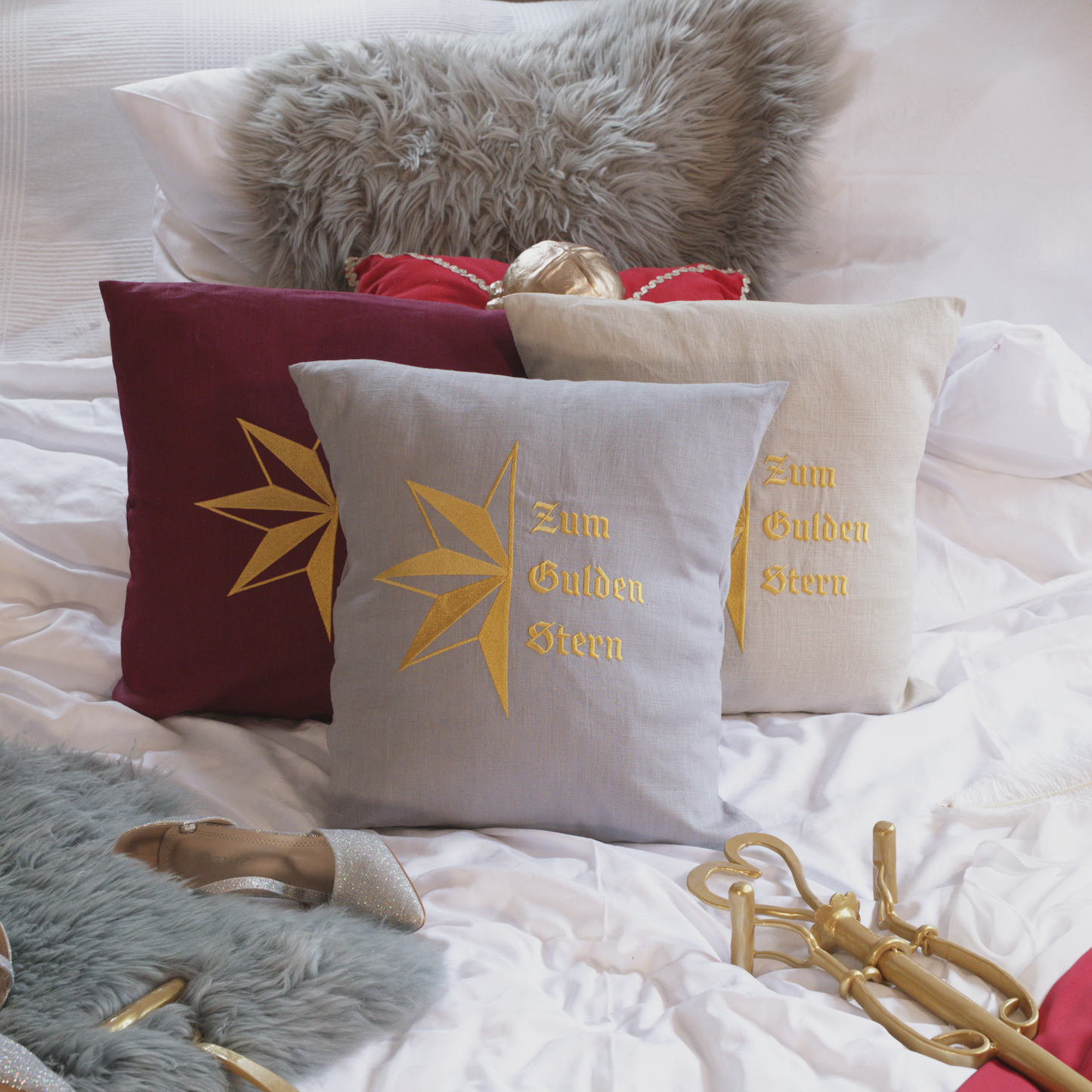 Drei dekorative Leinenkissen in den Farben Mondgrau, Sandstein und Weinrot mit goldener Stickerei, die den stilisierten Stern und die Aufschrift „Zum Gulden Stern“ zeigt. Die Kissen sind inklusive Füllung und liegen auf einem elegant arrangierten Bett mit weiteren Dekoelementen wie einem grauen Kunstfell-Kissen, einem roten Samtkissen mit goldenen Details, einem silbernen Schuh und einer goldenem Zepter.