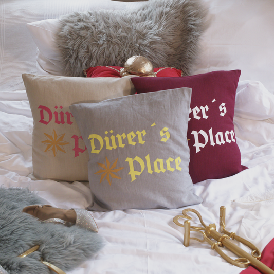 Drei dekorative Leinenkissen in den Farben Mondgrau, Sandstein und Weinrot mit kunstvoller Stickerei. Das Design zeigt die Aufschrift „Dürer’s Place“ in historischer Frakturschrift sowie ein stilisiertes Sternmotiv. Die Kissen sind inklusive Füllung und liegen auf einem geschmackvoll arrangierten Bett mit weiteren Dekoelementen wie einem grauen Kunstfell-Kissen, einem roten Samtkissen mit goldenen Details, einem silbernen Schuh und einem goldenen Zepter.