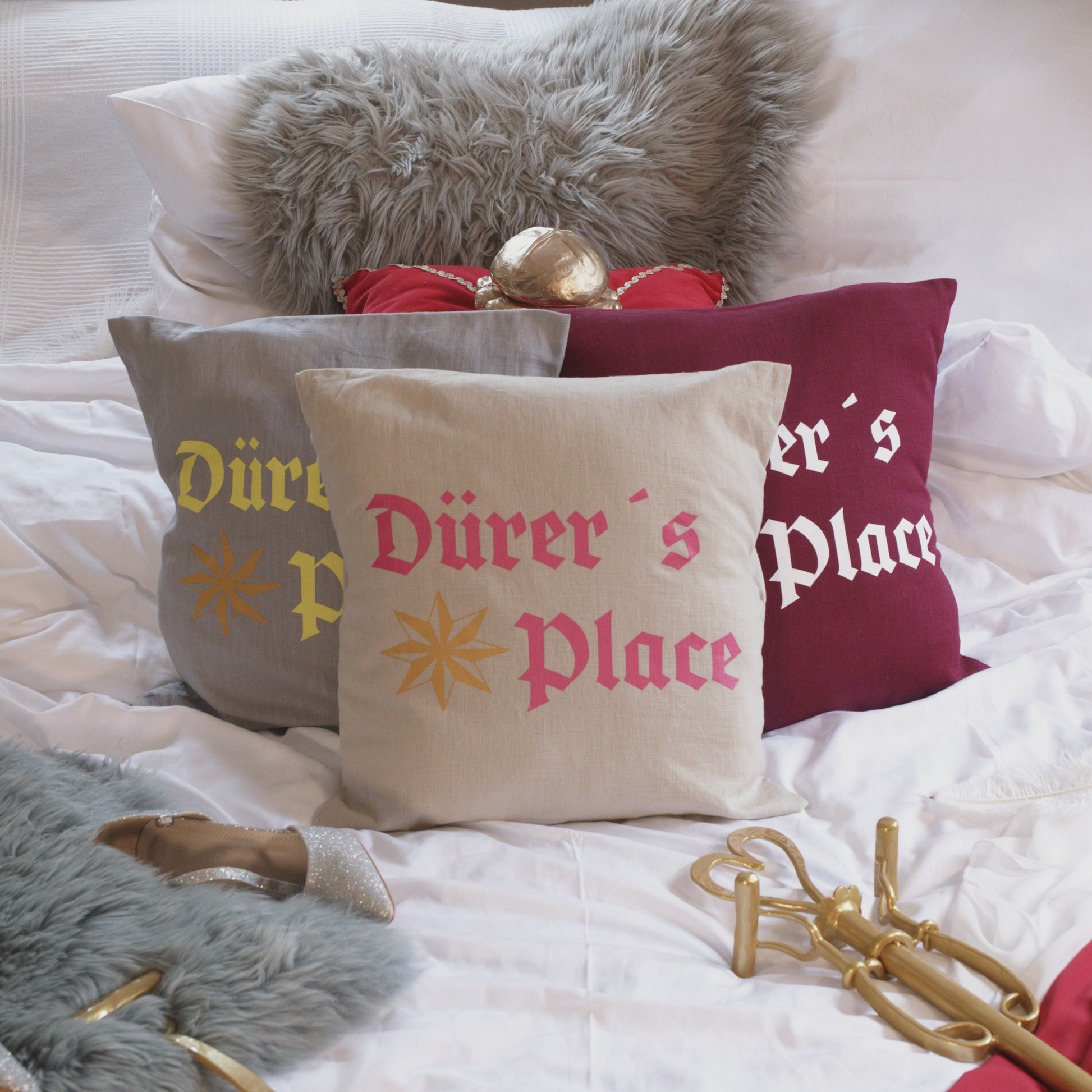 Drei dekorative Leinenkissen in den Farben Mondgrau, Sandstein und Weinrot mit kunstvoller Stickerei. Das Design zeigt die Aufschrift „Dürer’s Place“ in historischer Frakturschrift sowie ein stilisiertes Sternmotiv. Die Kissen sind inklusive Füllung und liegen auf einem geschmackvoll arrangierten Bett mit weiteren Dekoelementen wie einem grauen Kunstfell-Kissen, einem roten Samtkissen mit goldenen Details, einem silbernen Schuh und einem goldenen Zepter.
