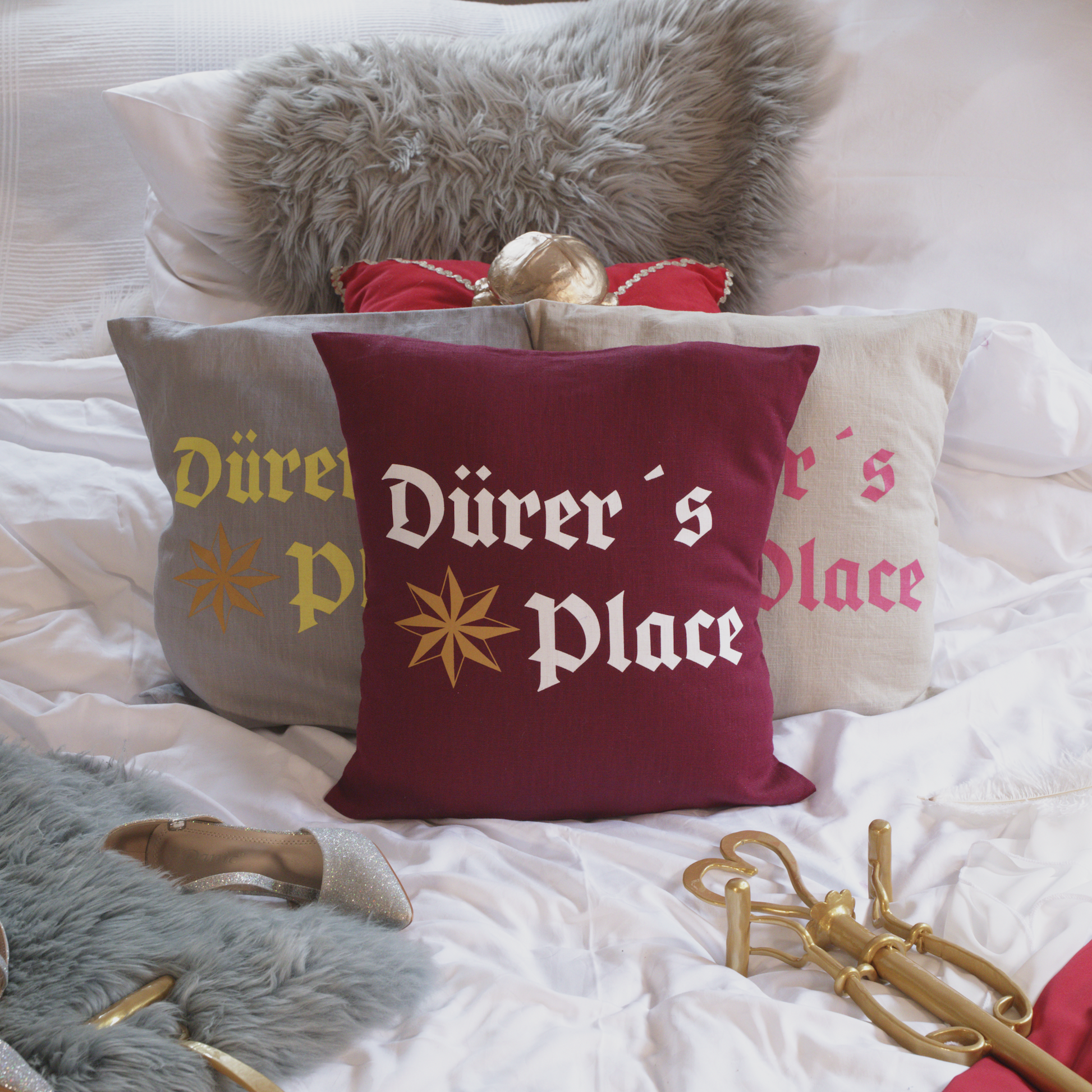 Drei dekorative Leinenkissen in den Farben Mondgrau, Sandstein und Weinrot mit kunstvoller Stickerei. Das Design zeigt die Aufschrift „Dürer’s Place“ in historischer Frakturschrift sowie ein stilisiertes Sternmotiv. Die Kissen sind inklusive Füllung und liegen auf einem geschmackvoll arrangierten Bett mit weiteren Dekoelementen wie einem grauen Kunstfell-Kissen, einem roten Samtkissen mit goldenen Details, einem silbernen Schuh und einem goldenen Zepter.