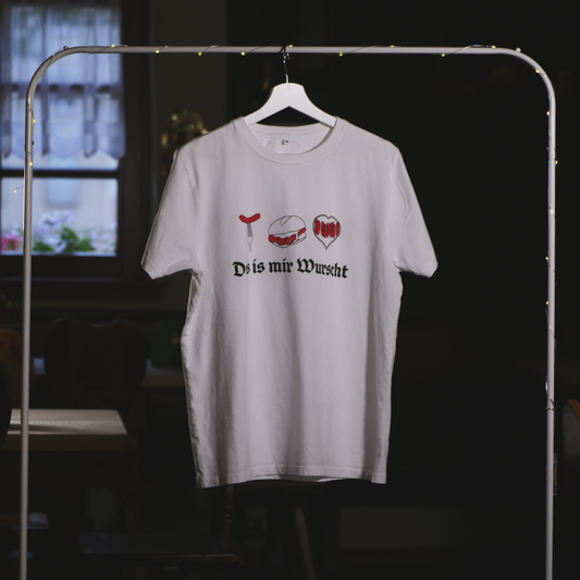 Ein weißes T-Shirt mit dem Aufdruck „Des is mir Wurscht“ hängt an einem weißen Kleiderbügel auf einem Kleiderständer mit einer dezenten Lichterkette. Das Design zeigt drei stilisierte Bratwurst-Motive – eine Bratwurst auf der Gabel, „Drei im Weggla“ und ein Bratwurst-Herz – kombiniert mit schwarzer Frakturschrift. Der Hintergrund ist eine gemütliche, rustikale Umgebung mit einem Fenster und traditionellen Holzmöbeln.