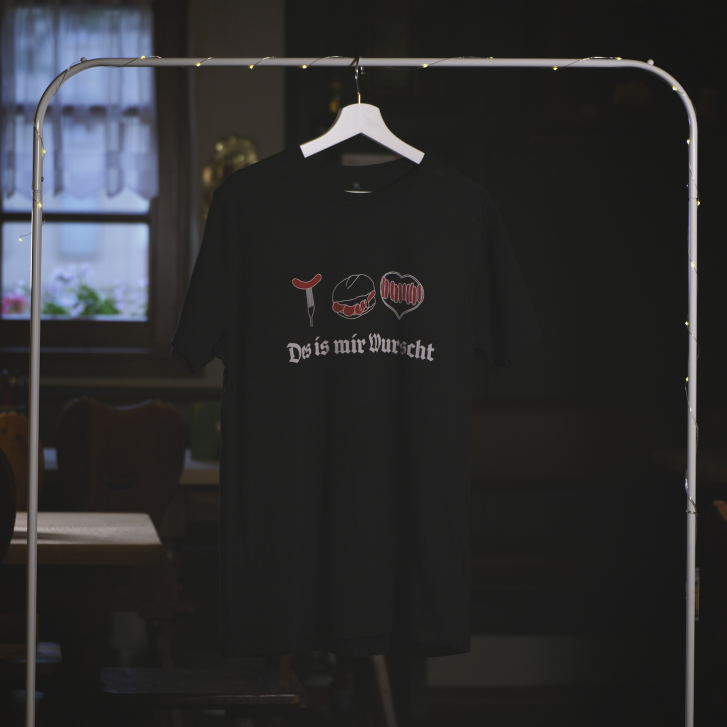 Ein schwarzes T-Shirt mit dem Aufdruck „Des is mir Wurscht“ hängt an einem weißen Kleiderbügel auf einem Kleiderständer mit Lichterkette. Das Design zeigt drei stilisierte Bratwurst-Motive – eine Bratwurst auf der Gabel, „Drei im Weggla“ und ein Bratwurst-Herz – in roten und weißen Akzenten, kombiniert mit schwarzer Frakturschrift. Der Hintergrund ist eine gemütliche, rustikale Umgebung mit einem Fenster und traditionellen Holzmöbeln.