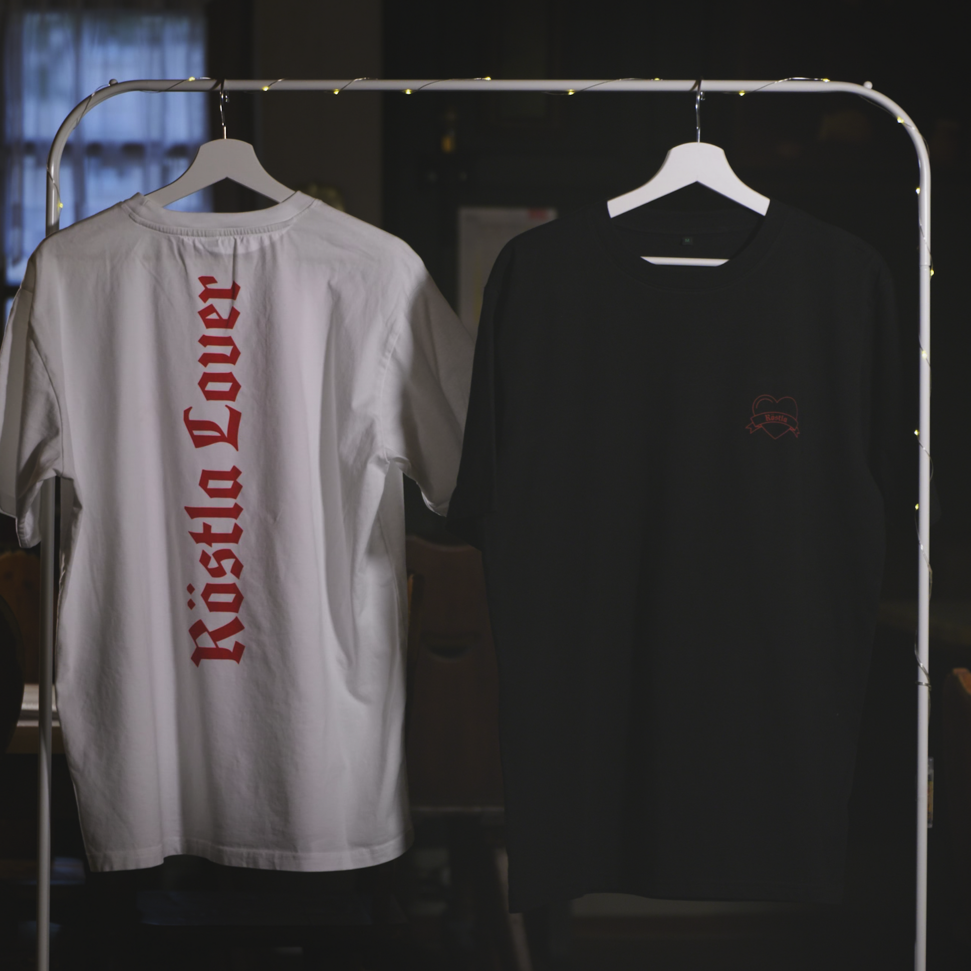 Zwei nachhaltige T-Shirts hängen an einem Kleiderständer mit Lichterkette. Das weiße Shirt zeigt auf der Rückseite die rote Aufschrift „Röstla Lover“ in gotischer Schrift. Das schwarze Shirt trägt auf der Vorderseite ein kleines, gesticktes Bratwurst-Herz mit der Aufschrift „Röstla“ in Rot. Beide Shirts bestehen aus 100 % Bio-Baumwolle, sind Fair Wear zertifiziert und klimaneutral produziert. Der Hintergrund zeigt eine gemütliche, rustikale Atmosphäre mit Fenstern und Holzmöbeln.