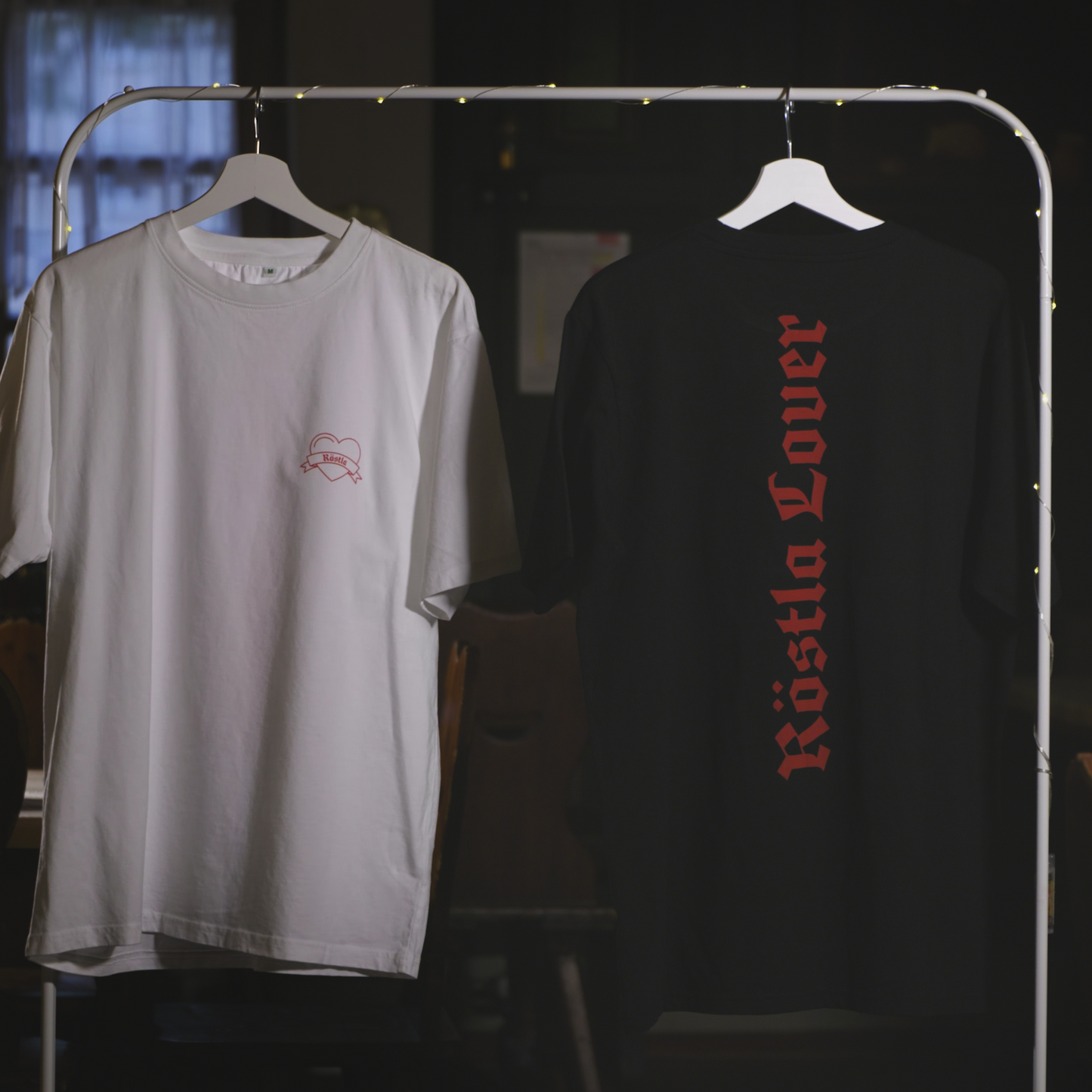 Zwei nachhaltige T-Shirts hängen an einem Kleiderständer mit Lichterkette. Das schwarze Shirt zeigt auf der Rückseite die rote Aufschrift „Röstla Lover“ in gotischer Schrift. Das weiße Shirt trägt auf der Vorderseite ein kleines, gesticktes Bratwurst-Herz mit der Aufschrift „Röstla“. Beide Shirts bestehen aus 100 % Bio-Baumwolle, sind Fair Wear zertifiziert und klimaneutral produziert. Der Hintergrund zeigt eine gemütliche, rustikale Atmosphäre mit Fenstern und Holzmöbeln.