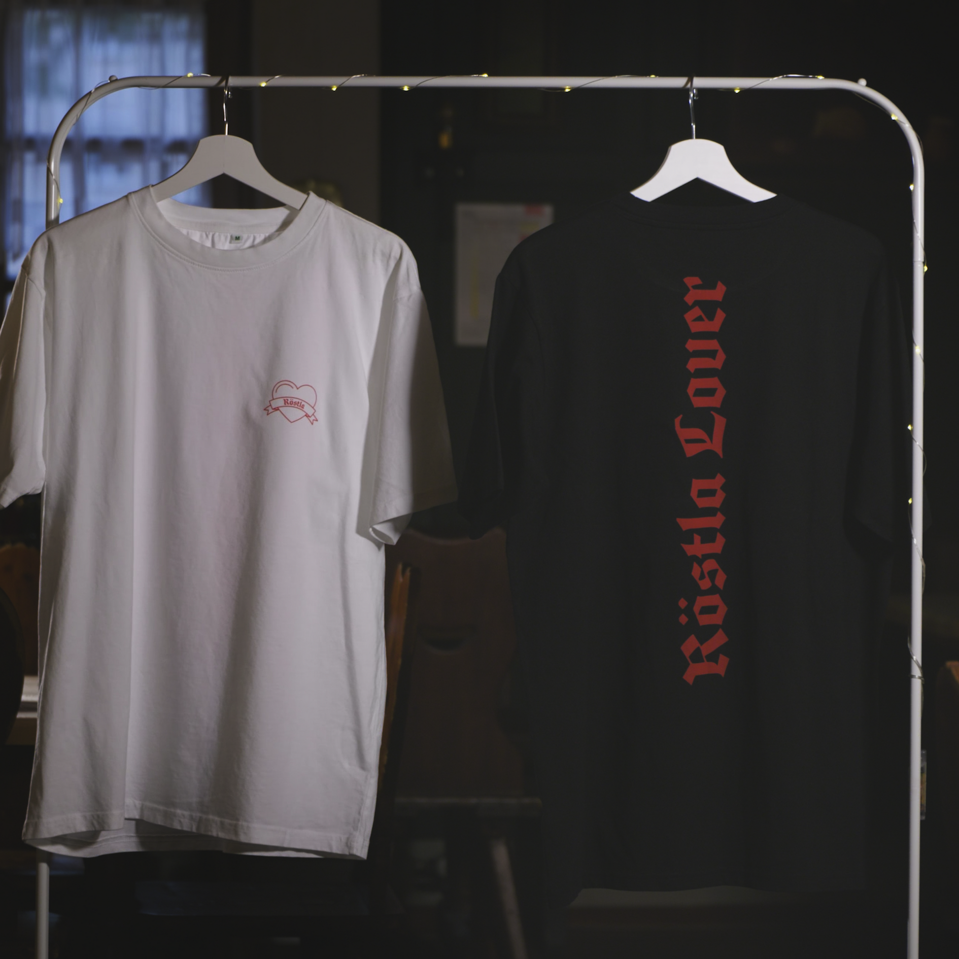 Zwei nachhaltige T-Shirts hängen an einem Kleiderständer mit Lichterkette. Das schwarze Shirt zeigt auf der Rückseite die rote Aufschrift „Röstla Lover“ in gotischer Schrift. Das weiße Shirt trägt auf der Vorderseite ein kleines, gesticktes Bratwurst-Herz mit der Aufschrift „Röstla“. Beide Shirts bestehen aus 100 % Bio-Baumwolle, sind Fair Wear zertifiziert und klimaneutral produziert. Der Hintergrund zeigt eine gemütliche, rustikale Atmosphäre mit Fenstern und Holzmöbeln.