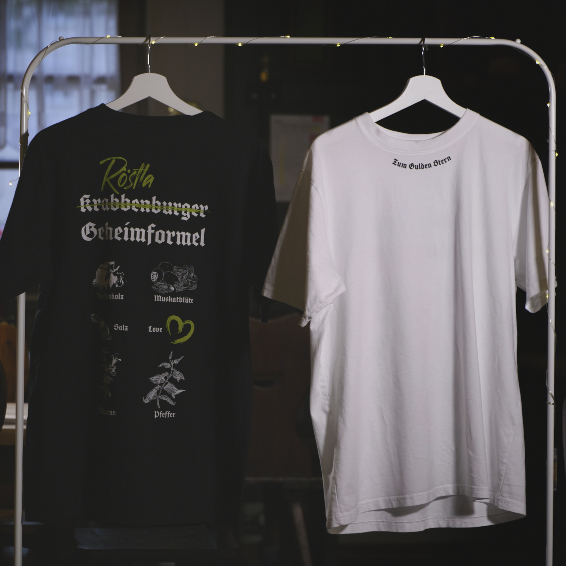 Zwei T-Shirts hängen an einem Kleiderständer mit Lichterkette. Das schwarze Shirt zeigt auf der Rückseite das Design „Röstla Krabbenburger Geheimformel“ mit grüner Schrift und illustrierten Zutaten wie Buchholz, Muskatblüte, Salz, Pfeffer und einem Herzsymbol für „Love“. Das weiße Shirt trägt auf der oberen Rückseite die dezente Aufschrift „Zum Gulden Stern“ in schwarzer Frakturschrift. Beide Shirts sind nachhaltig produziert, bestehen aus 100 % Bio-Baumwolle, sind Fair Wear zertifiziert und klimaneutral he