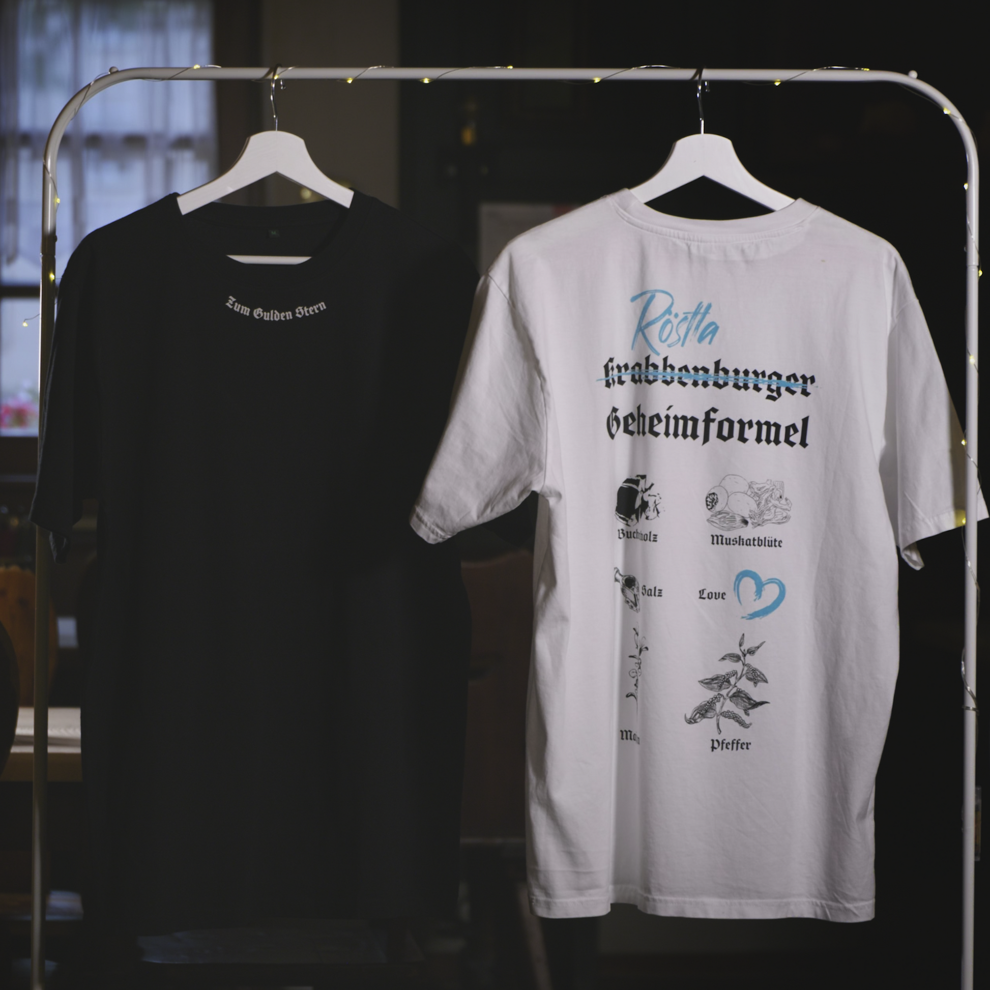 Zwei T-Shirts hängen an einem Kleiderständer mit Lichterkette. Das schwarze Shirt trägt auf der oberen Rückseite die Aufschrift „Zum Gulden Stern“ in weißer Frakturschrift. Das weiße Shirt zeigt auf der Rückseite das Design „Röstla Krabbenburger Geheimformel“ mit blauer und schwarzer Schrift sowie illustrierten Zutaten wie Buchholz, Muskatblüte, Salz, Pfeffer und einem Herzsymbol für „Love“. Beide Shirts bestehen aus 100 % Bio-Baumwolle, sind Fair Wear zertifiziert und klimaneutral produziert. Der Hintergru