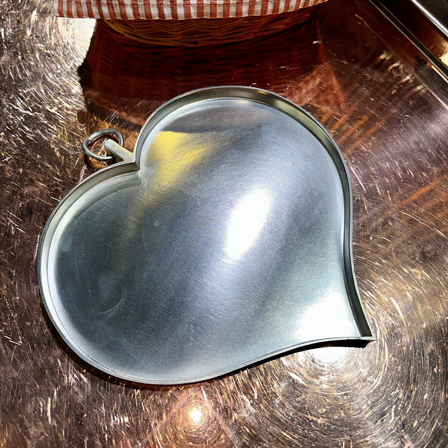 Ein herzförmiger Zinnteller mit gebürsteter Oberfläche liegt auf einer glänzenden Metallplatte. Die elegante Form mit sanften Rundungen und ein integrierter Aufhänger in Ringform machen den Teller vielseitig einsetzbar – sowohl als Servierplatte als auch als dekoratives Element. Lichtreflexe betonen die matte Struktur des hochwertigen Zinns.