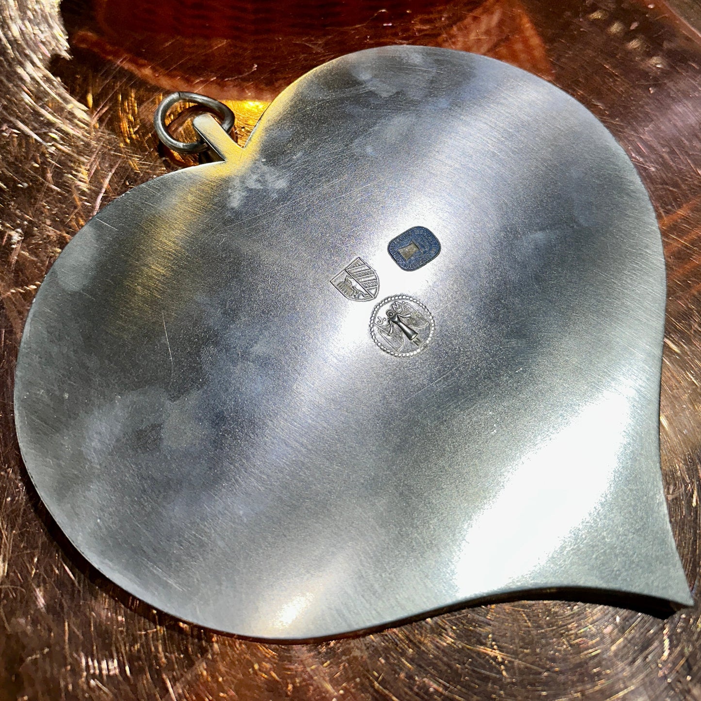 Ein herzförmiger Zinnteller mit einer gebürsteten, matten Oberfläche liegt auf einer glänzenden Metallplatte. Auf der Rückseite des Tellers sind mehrere Prägungen und Punzierungen sichtbar, darunter ein Wappen, eine stilisierte Figur und eine rechteckige Marke. Der Teller verfügt über einen integrierten Metall-Aufhänger in Ringform, wodurch er sowohl als Servierplatte als auch als dekoratives Element genutzt werden kann. Lichtreflexe betonen die Struktur des Zinns und verleihen dem Bild eine edle Anmutung.