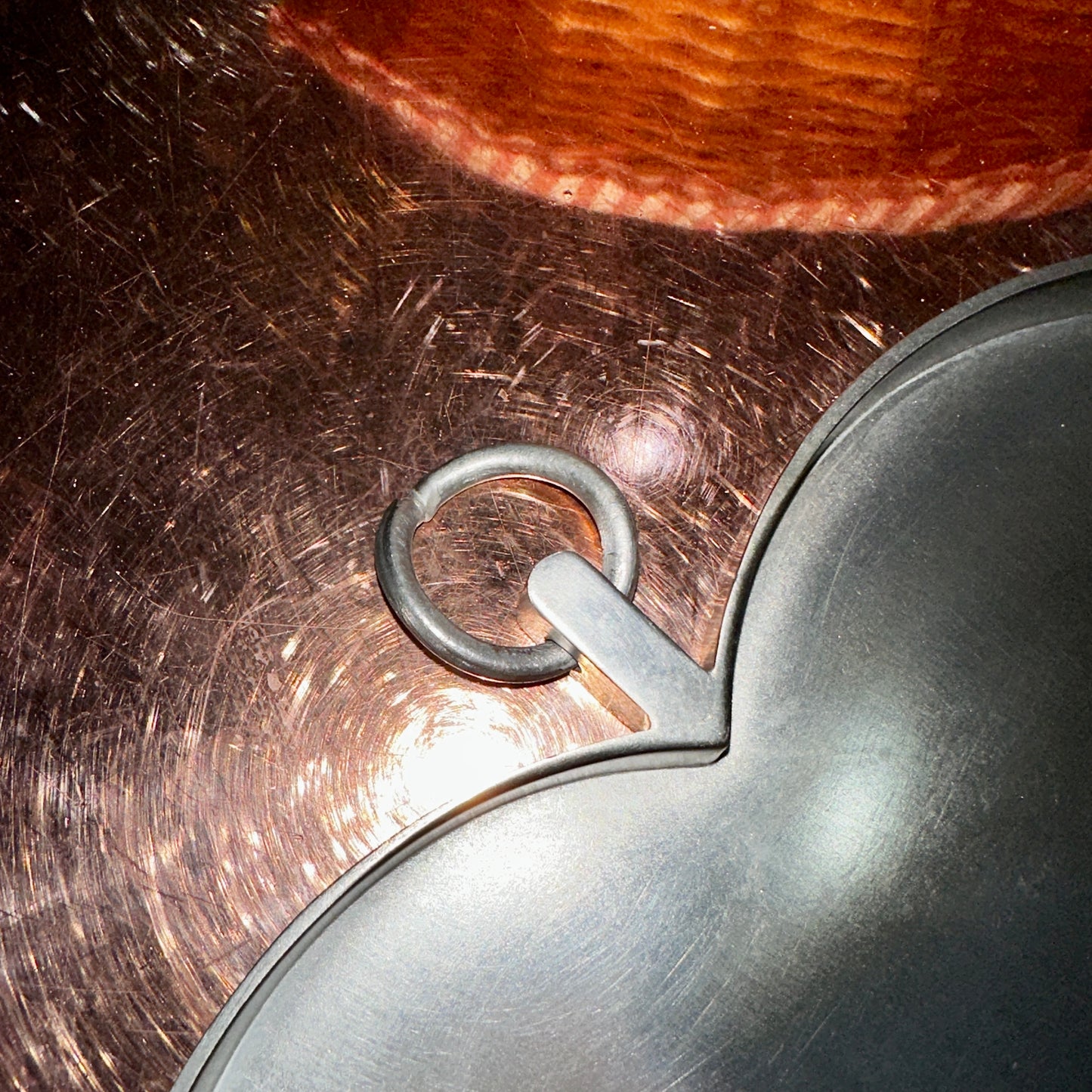 Nahaufnahme eines herzförmigen Zinntellers mit einem integrierten Metall-Aufhänger in Ringform. Der matte Zinn hat eine leicht gebürstete Oberfläche, die Lichtreflexe erzeugt. Der Hintergrund zeigt eine glänzende Metallplatte mit kreisförmigen Kratzspuren, die das Licht reflektiert. Die detaillierte Aufnahme hebt die handwerkliche Verarbeitung des Zinntellers hervor.