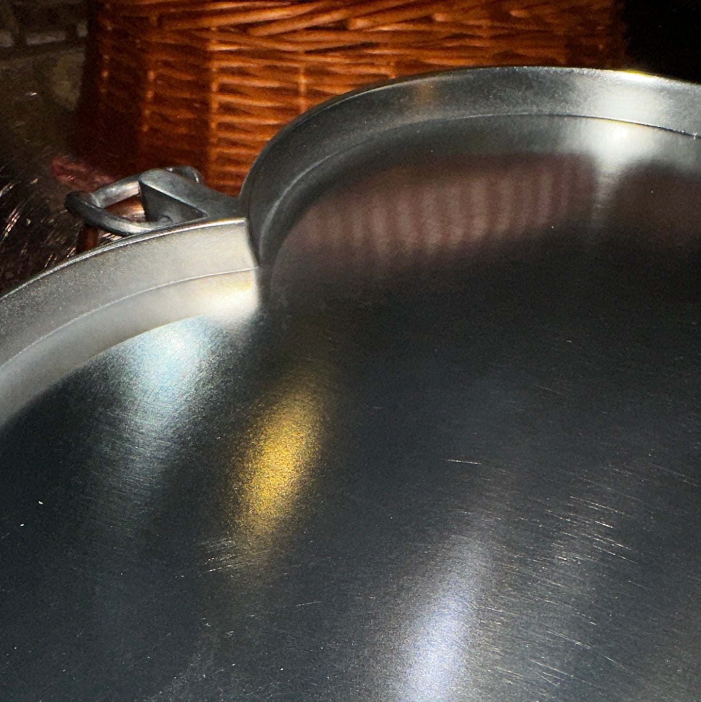 Nahaufnahme eines herzförmigen Zinntellers mit gebürsteter, matt glänzender Oberfläche. Lichtreflexe betonen die feine Struktur des Metalls, während der leicht erhöhte Rand und der integrierte Metall-Aufhänger in Ringform sichtbar sind. Der Hintergrund ist verschwommen, mit warmen Farbtönen, die sich sanft auf der reflektierenden Oberfläche spiegeln.