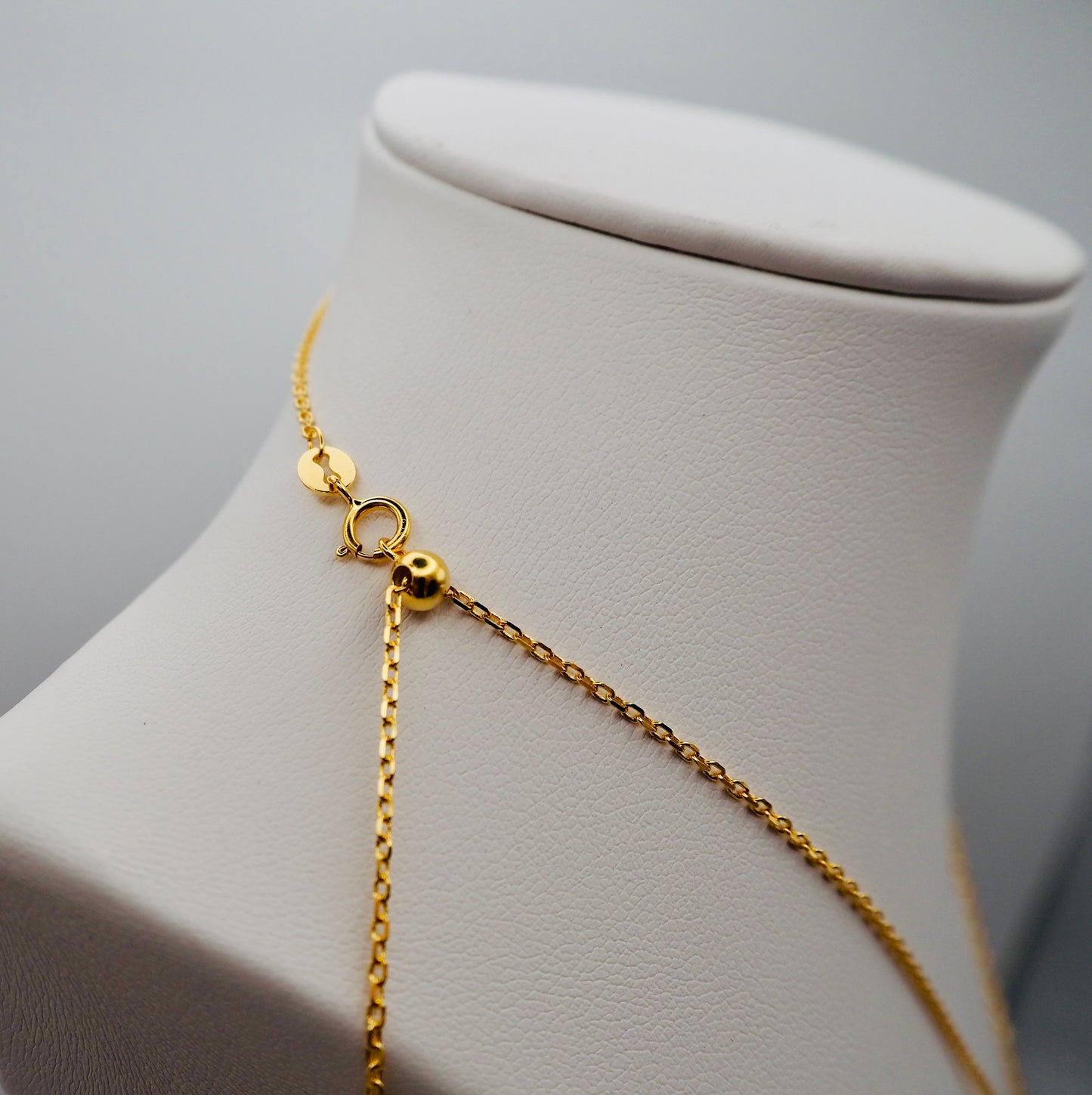 Nahaufnahme des Verschlusses einer eleganten, vergoldeten Kette, präsentiert auf einem weißen Schmuckdisplay. Die feingliedrige Kette verfügt über einen hochwertigen Karabinerverschluss und eine verstellbare Perle zur individuellen Längenanpassung. Ein kleines, rundes Plättchen am Verschluss sorgt für eine edle Detailveredelung.